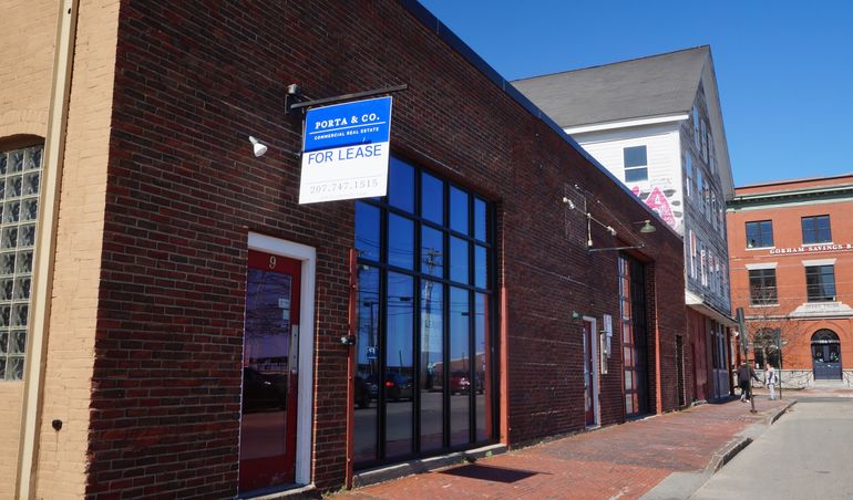 a one story brick building with a large window and a sign that says port & go for lease a brick sidewalk runs in front of it