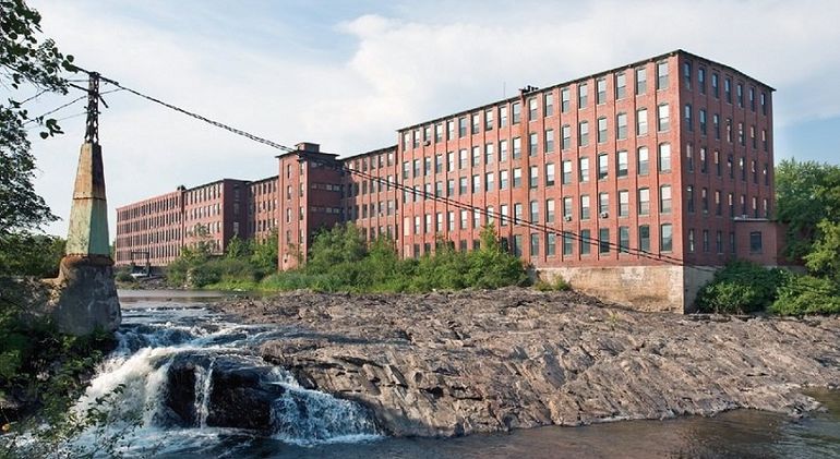 a large four-story brick 19th century mill building on the rocky edge of a river