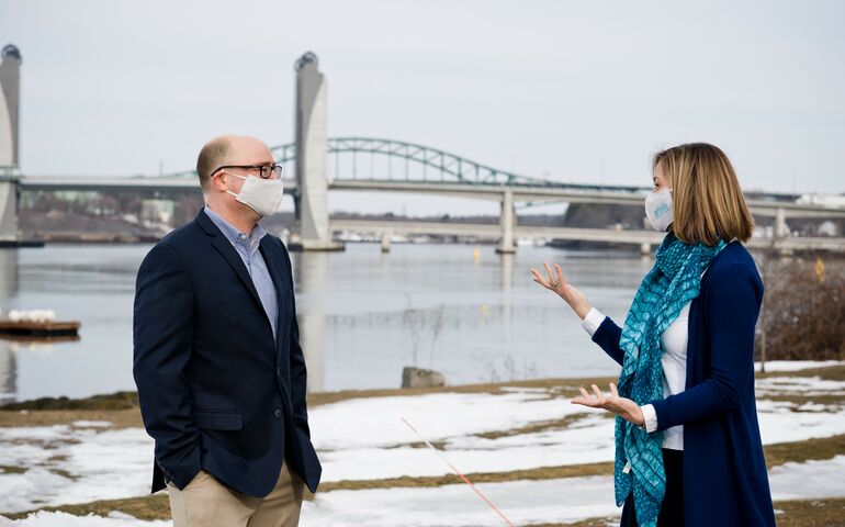 a white man and white woman in business attire and face coverings talk outdoors the ground is snowy and two large bridges stretch behind them