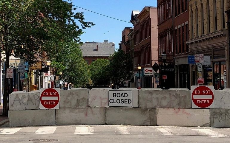 A concrete barricade with signs that say street closed do not enter blocks off a street with historic brick buildings and signs that show they are stores and restaurants
