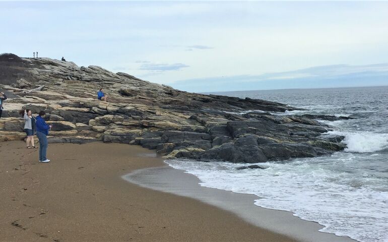 a beach with a large granite outcropping and a handful of people, both on the sand and up on the rocks watching frothy waves come in