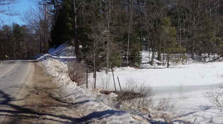 a stream goes under a country road, the opening has a small wire fence around it, it's winter and there's snow on the ground, and the stream is partially frozen and icy, pine trees line a ridge in the background