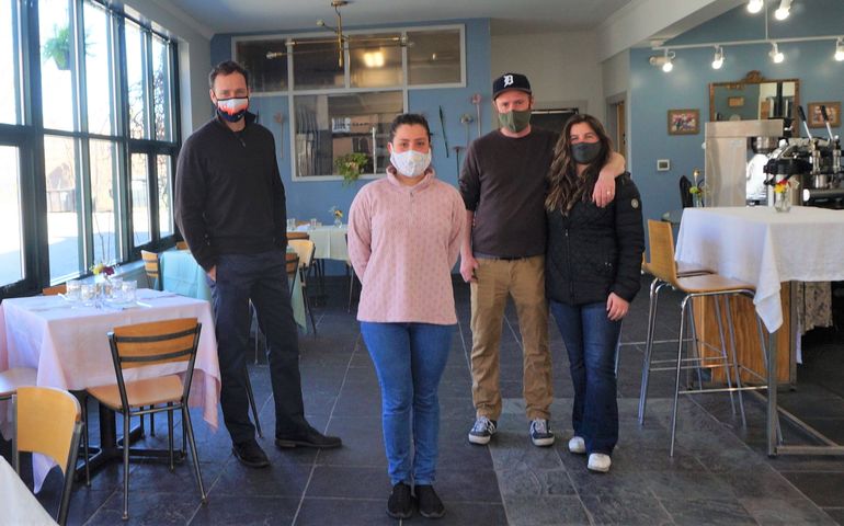 Four people in masks, a tall man, a smaller woman, another man, with a Detroit Tigers hat on and with his arm around the second woman, all white, stand in an empty restuarant that has big windows to the left and small tables with white table cloths