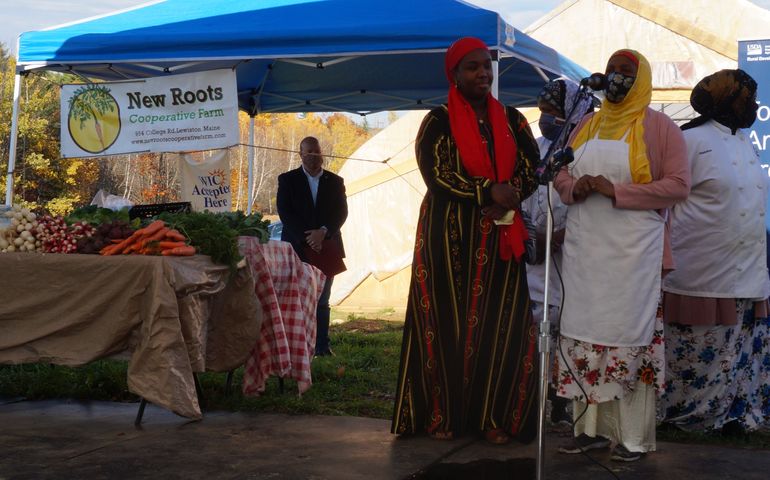 A Black woman dressed in traditional Somali clothing speaks at a microphone as others gather around her next to them is a table piled with produce and a sign that says New roots cooperative farm.