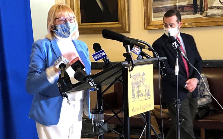 A woman, white, with short blond hair and wearing a face covering stands at a lectern that has microphones on it, a man in a suit, white, with dark hair and a face covering stands nearby a sign of the front of the lecturn says Maine legislature
