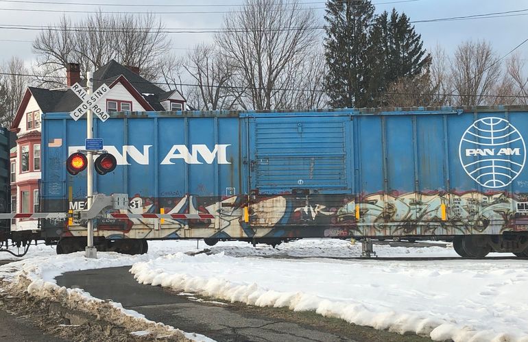 A blue train car with the words Pan Am crosses a road and sidewalk with a closed black and white striped railroad gate and flashing lights that say railroad crossing