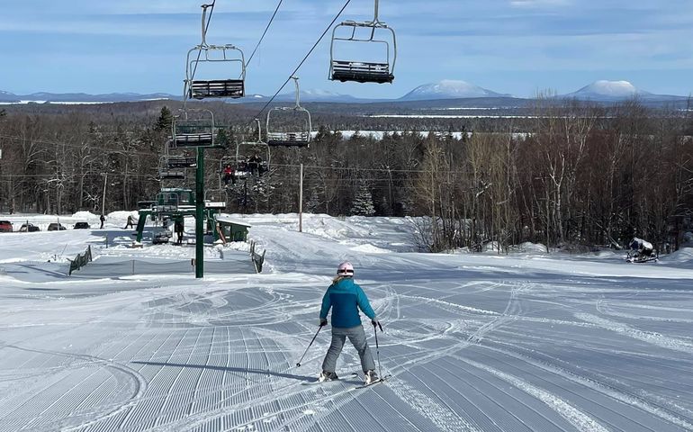 A ski lift with freshly groomed snow below with a skier on it and mountains in the background