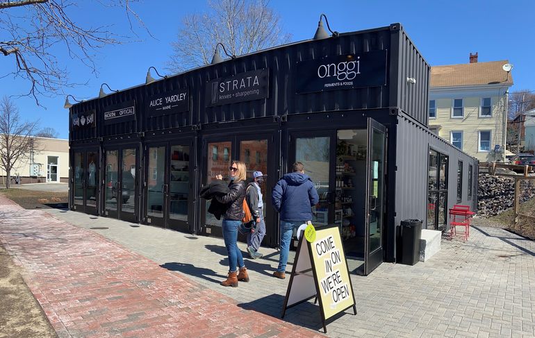 a small black building with five storefronts, the one at the close end has a sign over it that says onggi and a street sign that says come on in we're open as three white people mill about outside