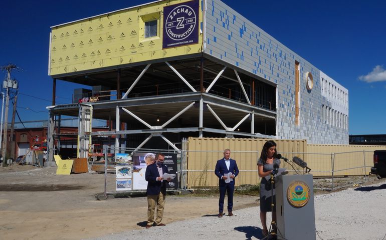 a woman, whit, with long dark hair reads at a podium in front of a large under construction cube shaped building as two white men in suits stand at a socially distanced distance behind her on what looks like a hot day