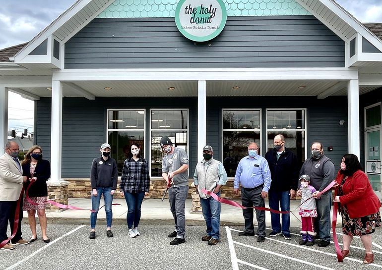 a large group cuts a ribbon in front of a building that says holy donut maine potato donuts