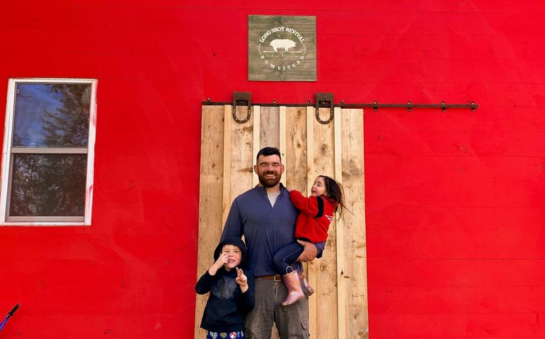 A man, white, with a beard, holds a little girl and has his arm around a little boy in front of a wooden barn door in a bright red barn with a sign long shot revival homestead above the door