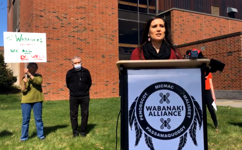 A woman, native american, stands at a lecturn with a sign that says wabanaki alliance as people holding signs supporting tribal sovereighnty stand behind her