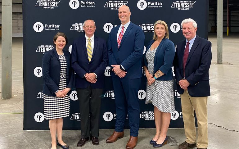 a group of five people, all white, two women and three men, standing in a large empty industrial room with a backdrop behind them that has logos for puritan medical products and tennessee department of economic and community development