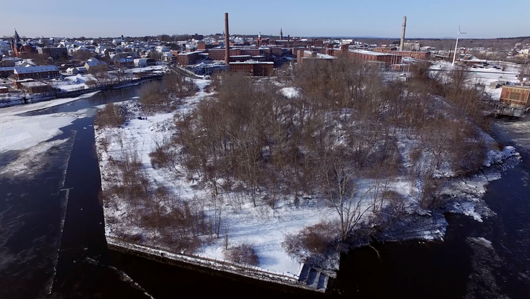 a vacant tract of land in a river, with snow on it, and behind it many brick mills