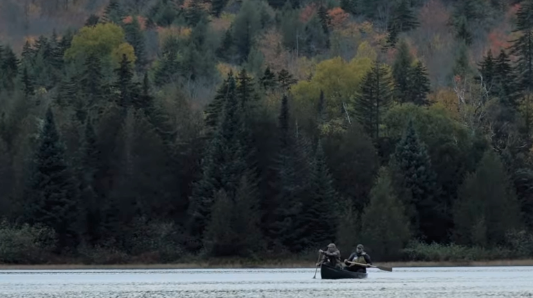 two men in a canoe early in the morning on placid water as evergreens rise behind them