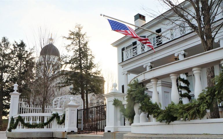 a white house with an ornate fence and american flag flying above the door in the foreground with a large granite building with a copper dome in the background