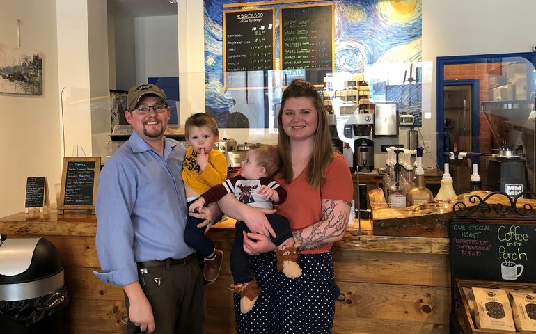 A young white couple each holds a little blond boy in front of a coffee counter that has signs for different blends and espressos