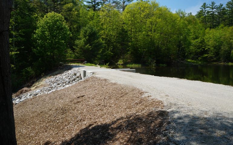 An earthen dam with a gravel road that goes over it is in a wooded area