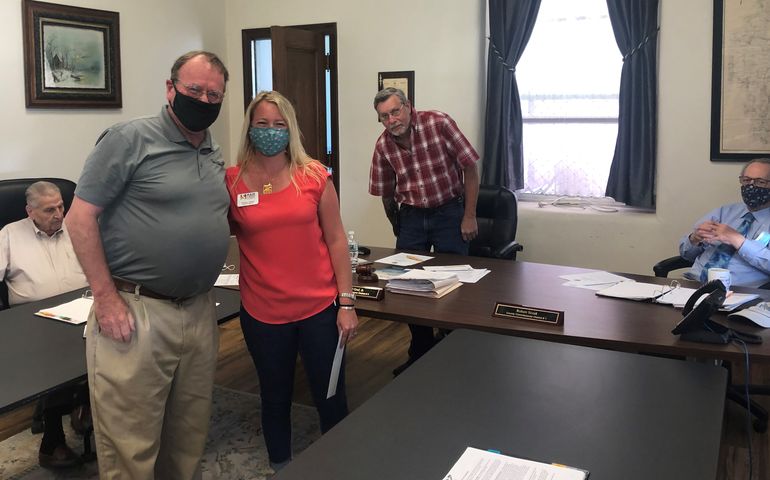 A woman in an office stands next to a man, both white and wearing facemasks, with three other men at a long table behind them