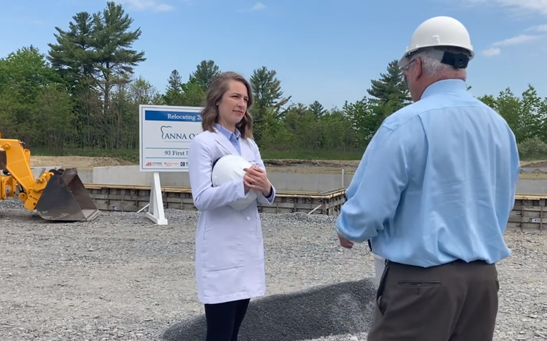 A white woman wearing a lab coat and holding a hard hat talks to a man, back to the camera, also white and wearing a heard hat at a gravel-covered constructin site with heavy equipment and a sign that says ouellet construction behind them.