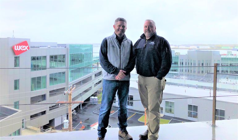 Two men, white, with beards, on top of a building with other modern buildings including one that says wex behind and below and the ocean in the background
