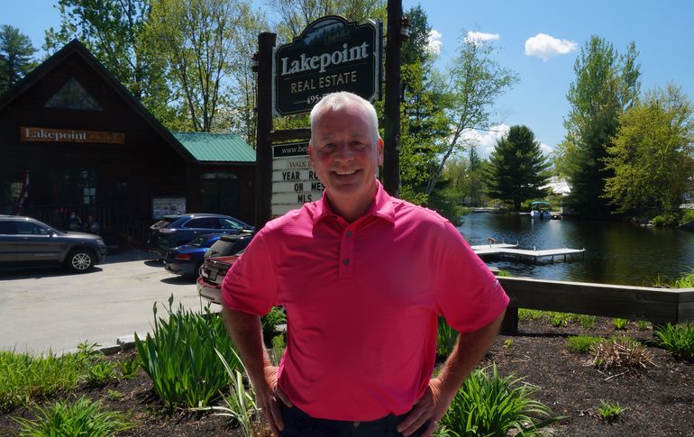 A smiling man, white with white hair, stands in front of a sign that says lakepoint with a water with docks behind him
