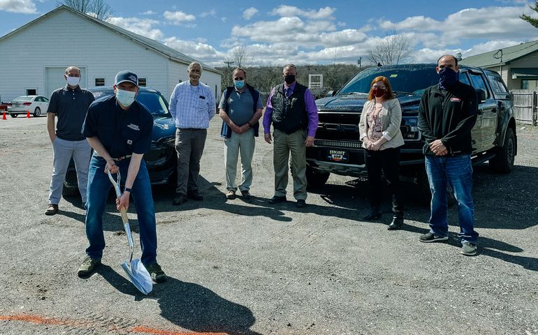 A man, white, with a face covering and dressed in work clothes, holds a shovel about a red line spray-painting across pavement as half a dozen people stand behind him 