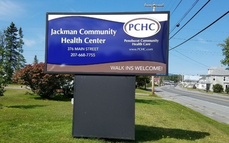 a sign that says jackman community health center pchc with a road in the background with wooden buildings