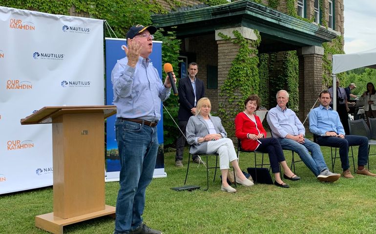 A man in a blue shirt jeans and baseball cap stands on grass holding a microphine and obviously saying something, as people nearby long on, including a tall bearded man standing and, seating in chairs. two women and two men, all white, behind them, part of a victorian three-story building is visible.