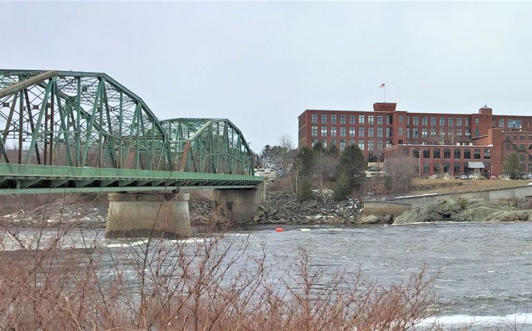 a green steel bridge over a river with a big brick mill on the other side