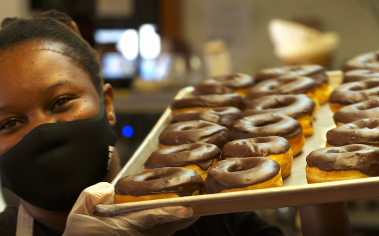 A woman wearing a face covering holds a tray of doughnuts