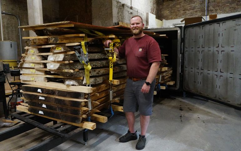 A man in t-shirt, shorts and with a beard stands next to a cart with a stack of large slabs of wood on it at the mouth of a large kiln