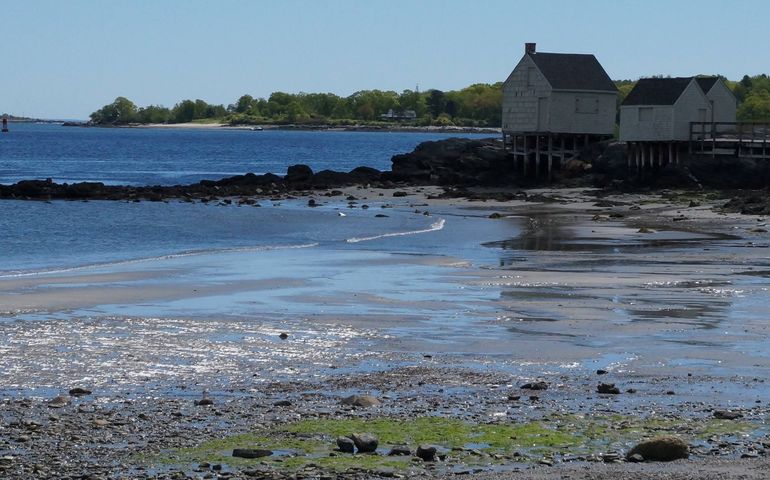 an old fishing shack on a rocky beach with the ocean spreading behind