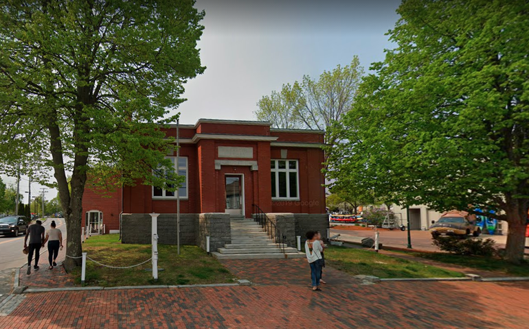 A small brick building with granite steps with a brick sidewalk in front, colorful kayaks can be seen leaning against the building next door