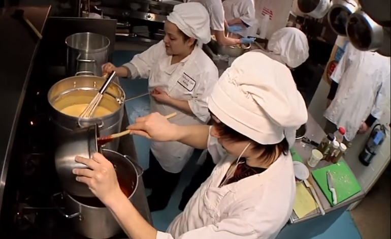 a young man and young women in chef's garb stir something in large pots