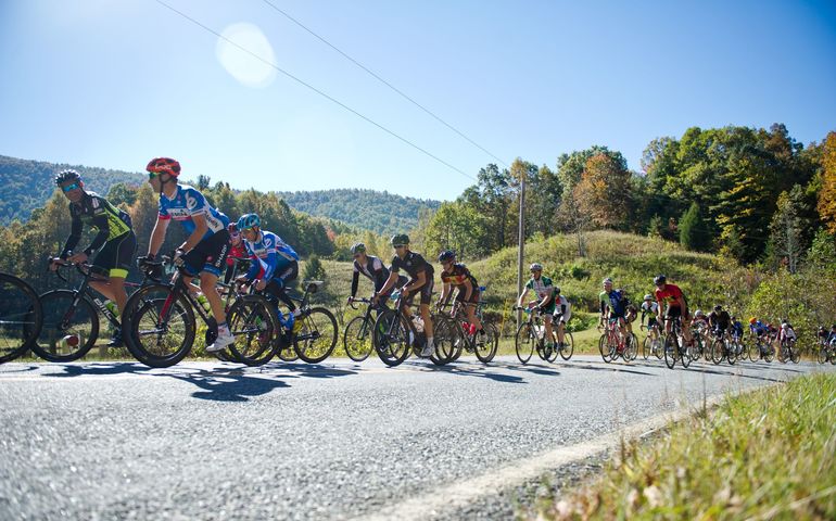 a pack of over a dozen competitive cyclists, cresting a hill surrounded by grass and trees