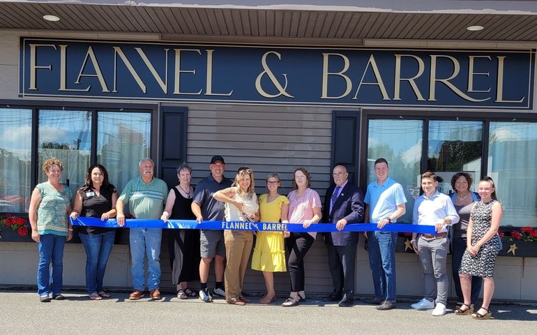 A group of people hold a sign in front of two large windows of a store, with flower boxes full of red flowers. A large sign reads FLANNEL & BARREL. A woman holds a large pair of scissors to a long ribbon for the store's ribbon cutting.