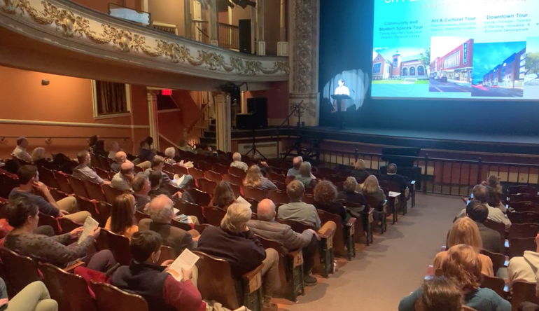 people seated in auditorium facing stage and screen