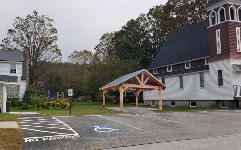 A wooden pavilion is seen between two buildings and set in a parking lot.