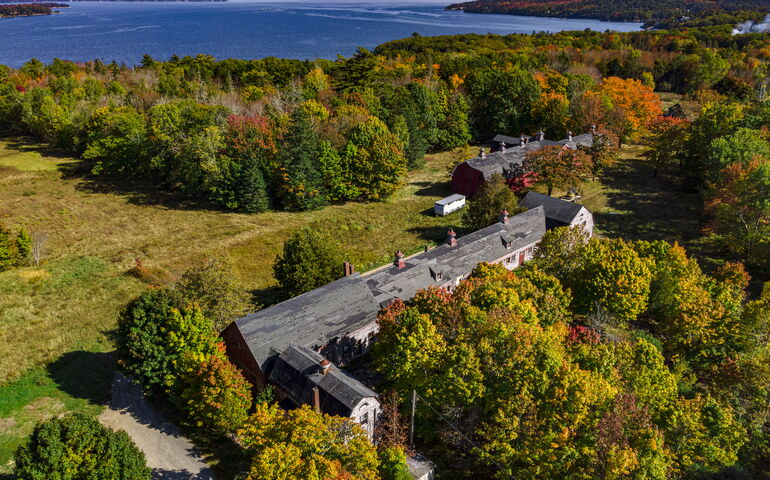 Aerial view of structures amidst woods and fields with a bay in the background.