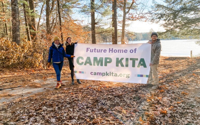 Three people hold a banner outside to advertise the new location of Camp Kita.