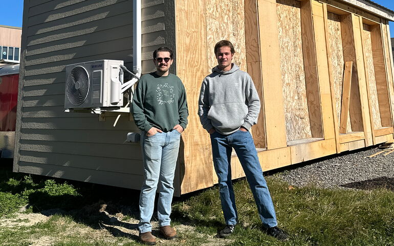 Two people stand in front of a wooden structure.