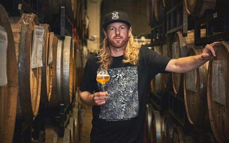 Tim Adams stands amidst wooden casks holding a glass of beverage.
