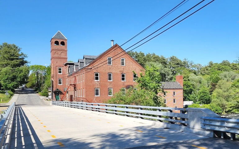 A old brick mill sits next to a road and bridge.