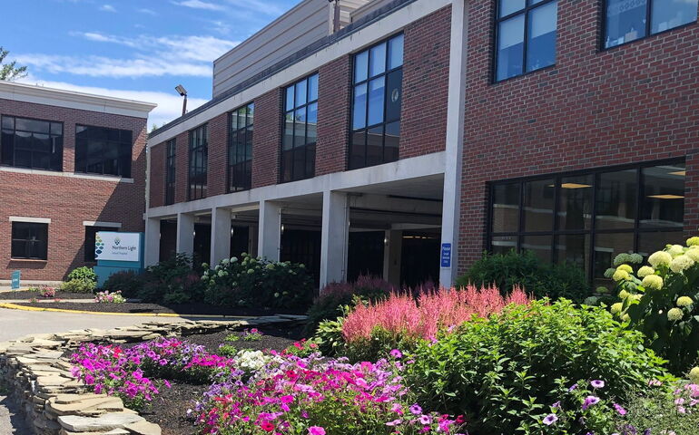 The exteiror of a brick and concrete building is fronted by flowering bushes.