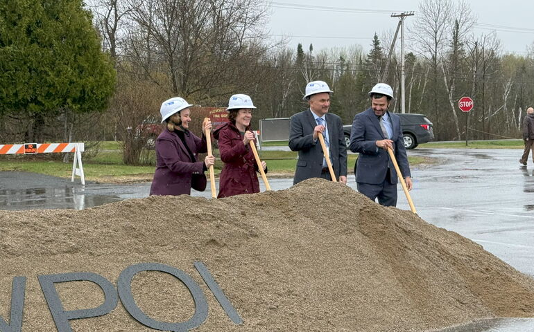 Four people stand in a row with shovels by a pile of sand.