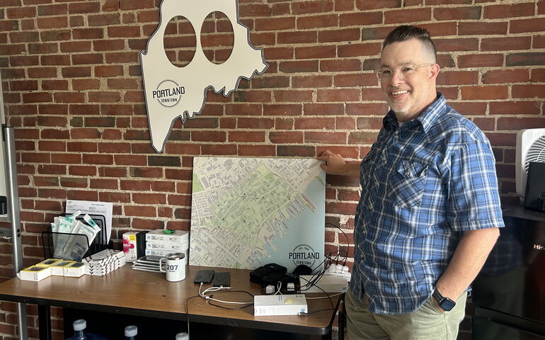 A person in a pliad shirts stands inside next to a table and hods a framed map.