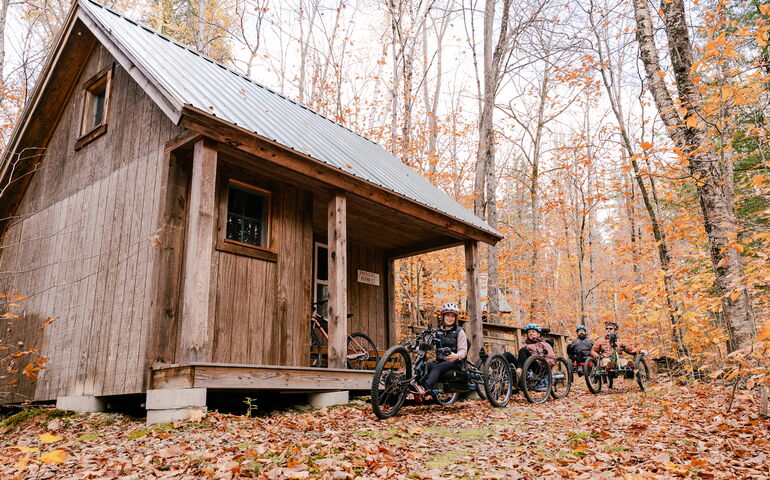 People on bikes ride through the woods past a cabin.