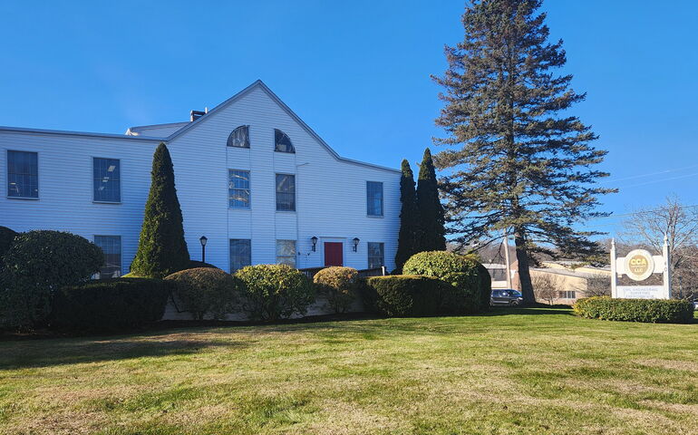 A building exterior has white siding, a sign, bushes and a lawn.