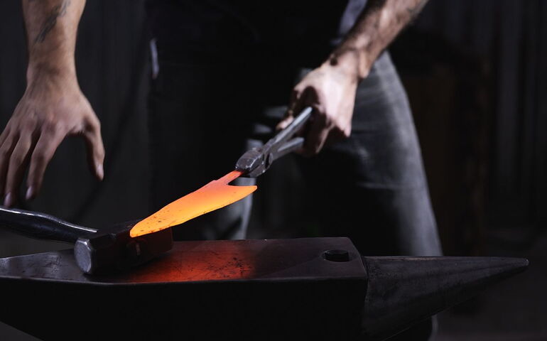 A person works at a forge and holds a red-hot piece of metal with a tool.